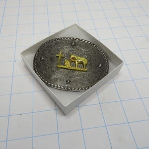 May include: Oval-shaped silver-tone belt buckle with a gold-tone cross and lamb design. The buckle is in a white box, with a decorative border and star accents. A religious-themed accessory.