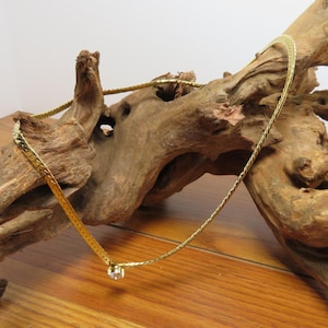May include: A gold chain necklace with a small, clear crystal pendant. The necklace is displayed on a piece of driftwood.