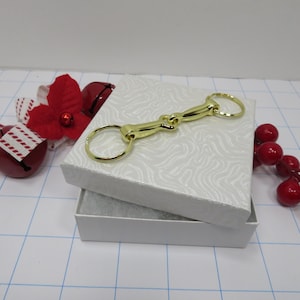 May include: A gold-colored metal horse bit keychain in a white gift box with a white and gray patterned lid. The box is open and the keychain is inside. The box is surrounded by red Christmas decorations.
