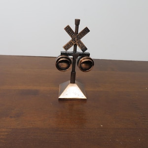 May include: A bronze-colored miniature railroad crossing sign with the words "RAIL CROSSING ROAD" on the sign. The sign has two round lights below the sign. The base is a square pyramid shape.