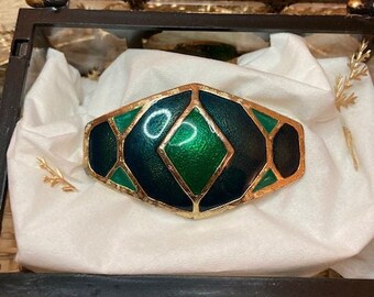 Könnte beinhalten: Ein breiter Armreif mit einem geometrischen Muster aus dunkelgrünen und hellen smaragdgrünen Emaille-Segmenten, die durch dünne, texturierte goldfarbene Metalllinien getrennt sind. Der Armreif hat eine zentrale Rautenform.
