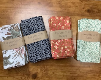 Puede incluir: Cuatro servilletas de tela plegadas con diferentes patrones florales y geométricos. Cada servilleta está envuelta con una banda de papel marrón que dice "Everyday Cloth Napkins". Los patrones incluyen diseños florales, geométricos y botánicos.