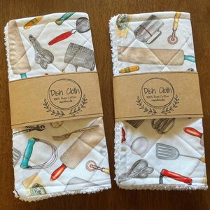 May include: Two white dish cloths with a quilted pattern and a variety of kitchen utensils printed on them. Each cloth is wrapped with a brown paper band that reads "Dish Cloth 100% Pure Cotton Handmade."