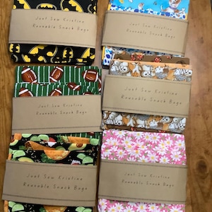 May include: A collection of reusable snack bags with various patterns, including a black and yellow bat symbol, footballs on green, and pink daisies. Each bag is wrapped with a brown paper band that reads "Just Sew Kristine Reusable Snack Bags."