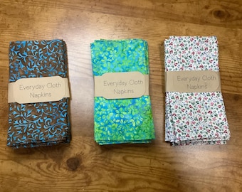 Puede incluir: Tres servilletas de tela plegadas con diseños decorativos. Una es marrón y azul, otra verde y azul, y la tercera blanca con flores rojas y rosas. Cada servilleta lleva la etiqueta "Everyday Cloth Napkins".