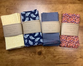Puede incluir: Cuatro pilas de servilletas de tela en varios patrones y colores. Una pila es amarilla con un diseño floral, otra es azul marino con paisley blanco, una tercera es azul con un pequeño patrón de puntos y la última es roja con un patrón floral. Cada pila está envuelta con una banda de papel marrón que dice "Just Sew Kristina Everyday Cloth Napkins".