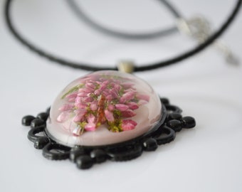 May include: A delicate necklace with a black cord and a pendant featuring a pink flower pressed inside clear resin. The pendant is set in a black metal frame with a decorative design.