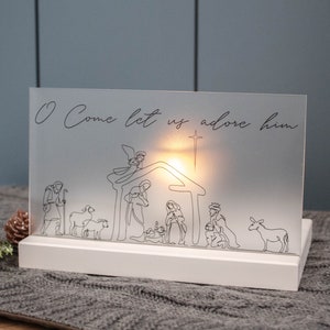 A white rectangle base sits on gray table runner, the base has a slot acting as a stand for a frosted acrylic piece.  The acrylic has a hand drawn nativity in black ink and allows light from a candle behind to filter through like a luminary.