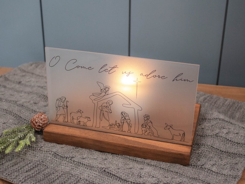 Walnut stained wood base sits on a gray table. The wood base acts as a stand for a frosted acrylic piece with a line drawn nativity printed in black on it. A candle sits in the stand behind the acrylic and illuminates the design.