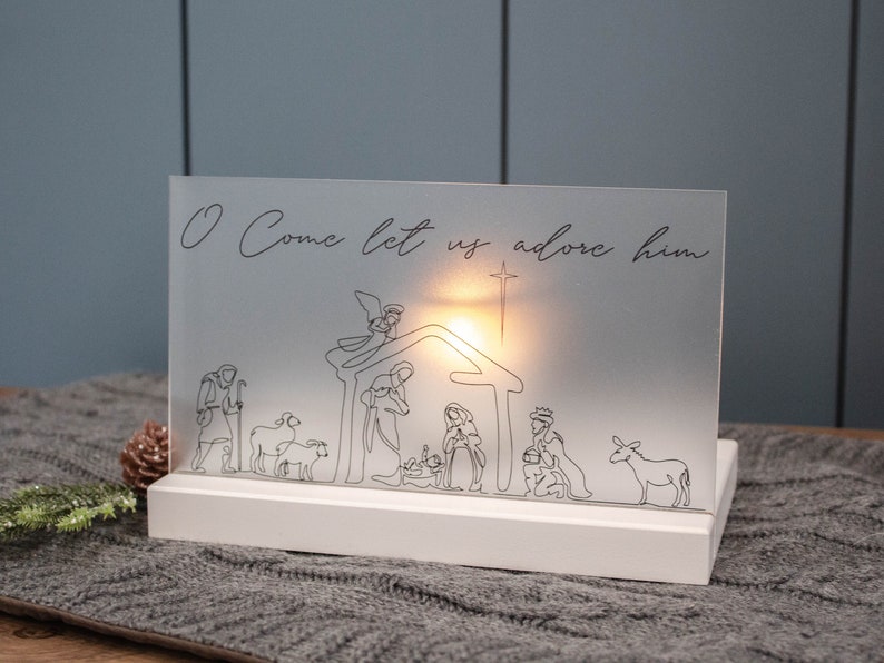 A white rectangle base sits on gray table runner, the base has a slot acting as a stand for a frosted acrylic piece.  The acrylic has a hand drawn nativity in black ink and allows light from a candle behind to filter through like a luminary.