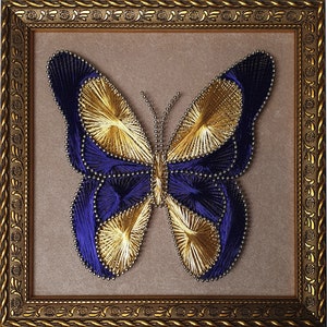 Könnte beinhalten: Gerahmtes String-Art-Schmetterling. Der Schmetterling besteht aus marineblauem und goldenem Faden, mit einem perlenbesetzten Körper und Fühlern. Der goldene Rahmen hat detaillierte Verzierungen. Das Kunstwerk wird vor einem neutralen Hintergrund präsentiert.
