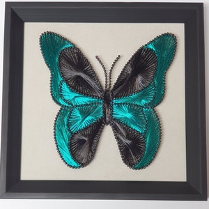 Op de afbeelding: Een ingelijst kunstwerk met een turquoise en zwarte vlinder gemaakt van string art. De vlinder is gecentreerd op een beige canvas en ingelijst in een zwart frame.