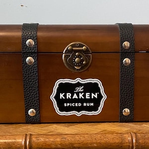 May include: A wooden chest with a dark brown finish, black leather straps, and silver studs. The chest features a brass clasp and a label that reads "The KRAKEN SPICED RUM."