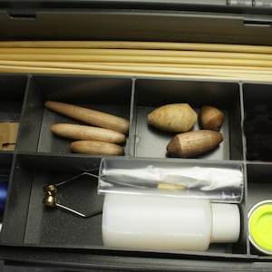 May include: A black plastic tackle box with compartments containing fly-fishing materials. It includes wooden dowels, wooden shapes, black material, blue thread, a fly-tying tool, a bottle, and two small containers.