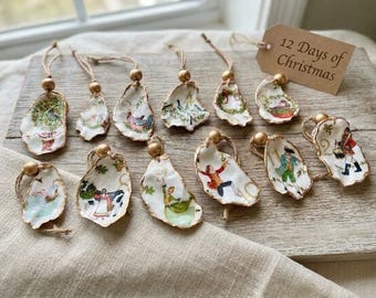 May include: Twelve oyster shell ornaments featuring illustrations representing the 12 Days of Christmas, each with a gold bead and twine loop. A tan paper tag with the text 12 Days of Christmas is included with the set of decorative shells.