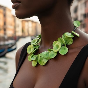Puede incluir: Una mujer con un collar verde vibrante compuesto por colgantes circulares en forma de hoja. El collar destaca sobre un top negro, con un fondo borroso de un canal y edificios. El collar es una pieza de joyería llamativa.