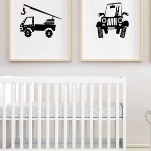 May include: Two black and white illustrations of vehicles, a crane truck and a jeep, are framed and hung on a white wall above a white cot with a grey polka dot sheet.