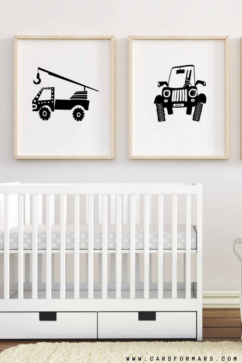 May include: Two black and white illustrations of vehicles, a crane truck and a jeep, are framed and hung on a white wall above a white cot with a grey polka dot sheet.