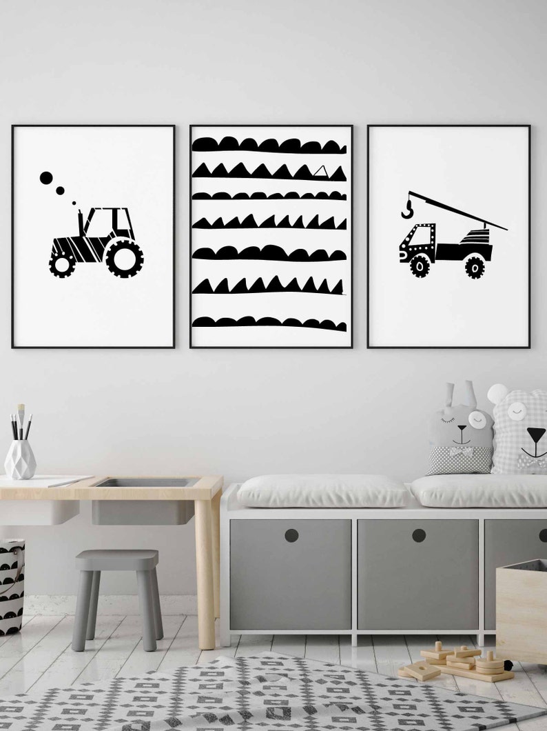 May include: Three black and white framed prints featuring a tractor, a truck with a crane, and a geometric pattern of waves and triangles. The prints are hanging on a white wall above a wooden desk and a grey storage bench with cushions.