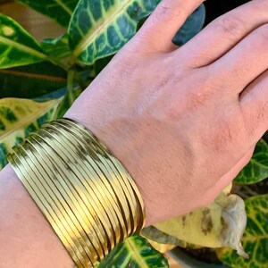 May include: A gold-colored cuff bracelet with a layered design. The bracelet is worn on a wrist, with green and yellow foliage in the background. The bracelet has a shiny, metallic finish and appears to be made of metal.