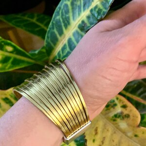 May include: A wide, gold-colored cuff bracelet with a series of parallel, slightly curved bands. The bracelet has a rectangular silver-colored clasp. The background features green and yellow leaves.