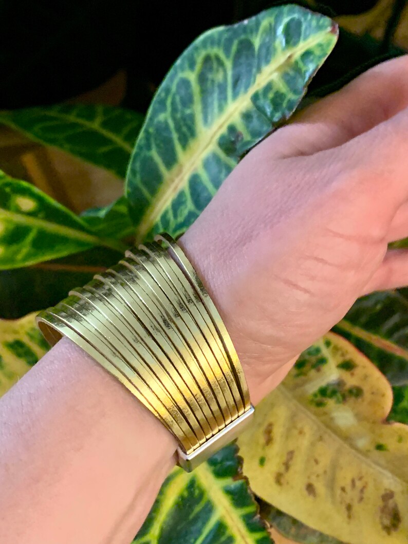 May include: A wide, gold-colored cuff bracelet with a series of parallel, slightly curved bands. The bracelet has a rectangular silver-colored clasp. The background features green and yellow leaves.
