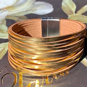May include: A multi-strand bracelet with a magnetic clasp. The bracelet features multiple layers of thin, gold-colored bands stacked on top of each other. The inner band is a light brown color. The clasp is silver.