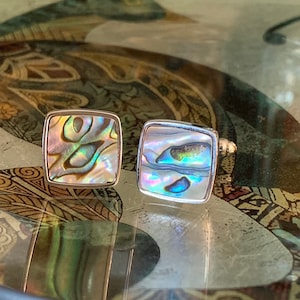May include: Two square cufflinks with iridescent abalone shell inlays set in silver. The abalone shells display a range of colors, including blues, greens, and purples. The cufflinks are displayed on a decorative surface.