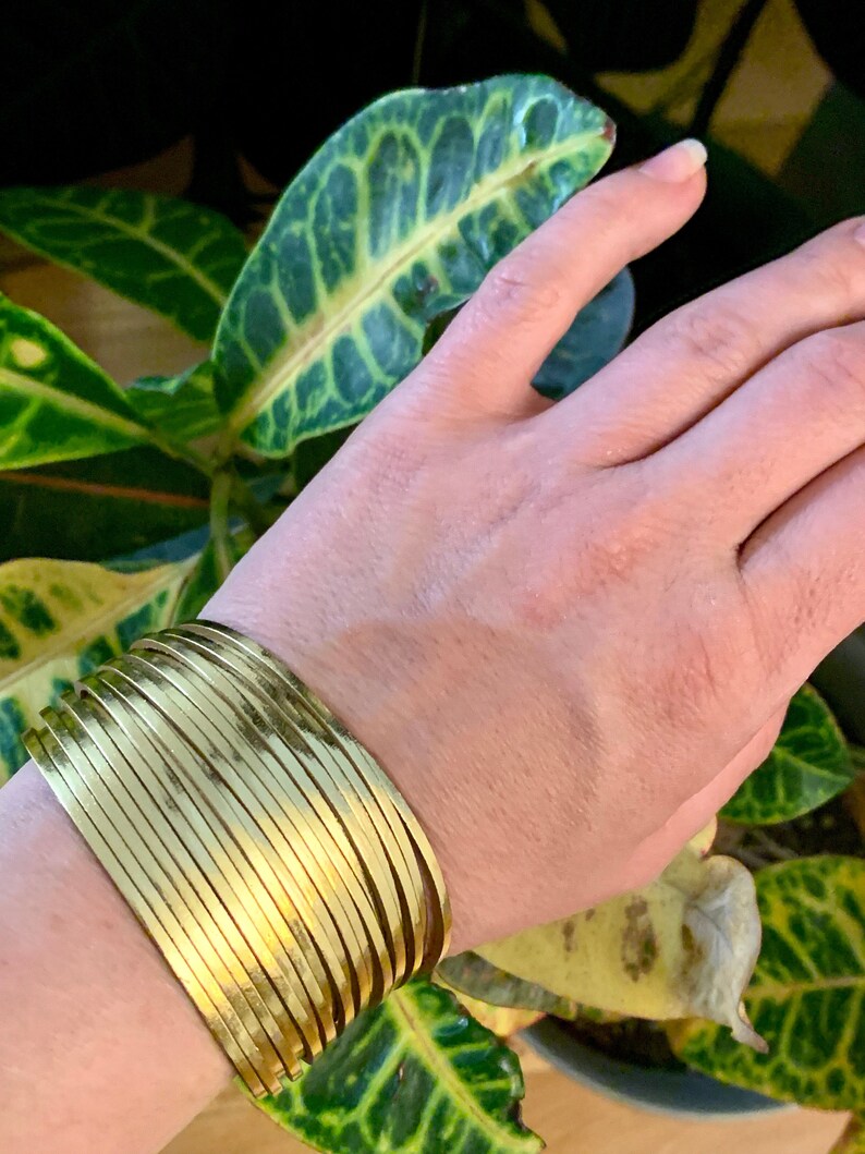 May include: A gold-colored cuff bracelet with a layered design. The bracelet is worn on a wrist, with green and yellow foliage in the background. The bracelet has a shiny, metallic finish and appears to be made of metal.