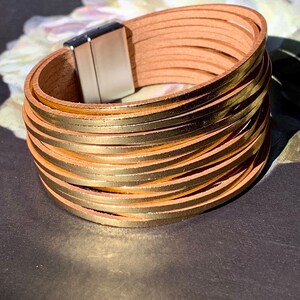 May include: A multi-strand bracelet with tan leather straps and gold-colored metal accents. The bracelet has a magnetic clasp and is designed to wrap around the wrist multiple times. The bracelet is a fashion accessory.