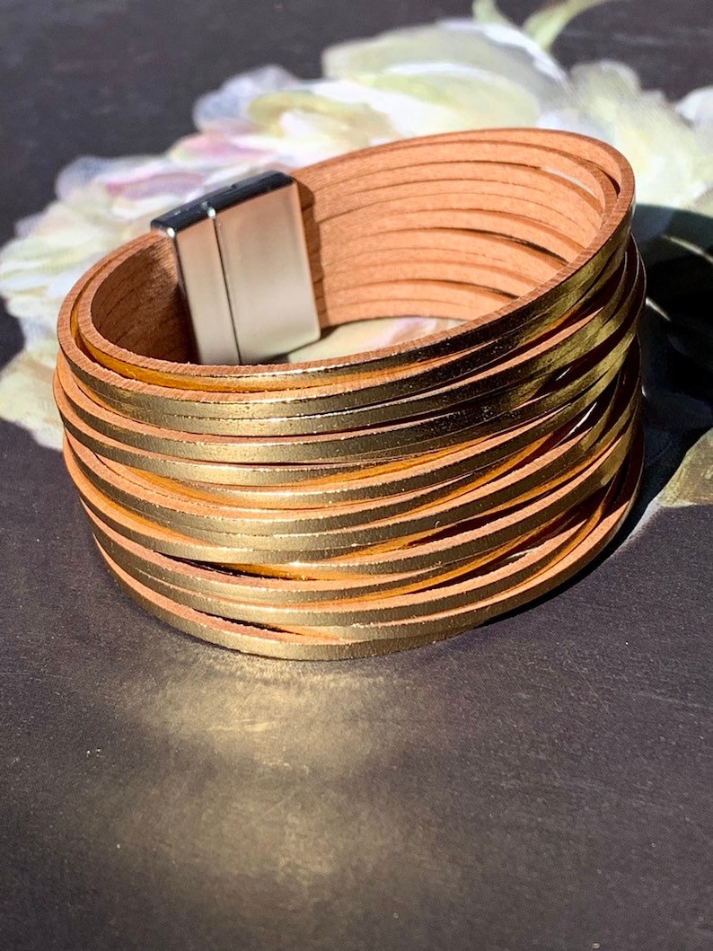 May include: A multi-strand bracelet with tan leather straps and gold-colored metal accents. The bracelet has a magnetic clasp and is designed to wrap around the wrist multiple times. The bracelet is a fashion accessory.