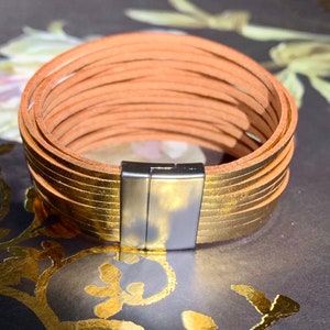 May include: A multi-strand bracelet with a magnetic clasp. The bracelet features multiple strands of tan leather and gold-colored leather. The clasp is silver-colored. The bracelet is approximately 7 inches in circumference.