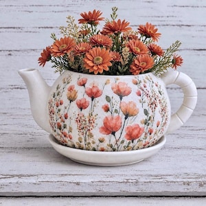 May include: A white ceramic teapot-shaped planter with a matching saucer, decorated with a delicate watercolor-style print of orange and pink wildflowers. The planter is filled with a bouquet of vibrant orange artificial daisy-like flowers.