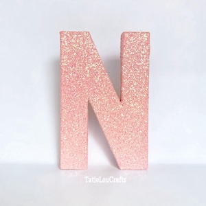May include: A large, free-standing letter "N" in a soft pink hue, covered in sparkling glitter. The letter is displayed against a plain white background. The Etsy shop name "TatieLouCrafts" is visible at the bottom.