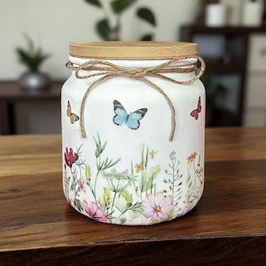 May include: A white jar with a wooden lid and twine bow. The jar is decorated with colorful butterflies and floral designs. The jar is sitting on a wooden surface.