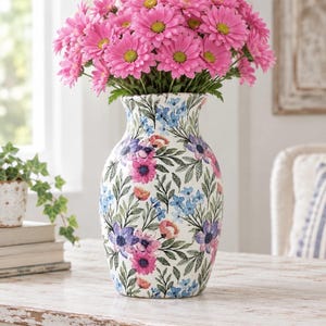 May include: A white ceramic vase with a tapered neck and a rounded body, decorated with a repeating pattern of pink, purple, and blue flowers with green leaves. The vase is filled with a large bouquet of vibrant pink daisy-like flowers.