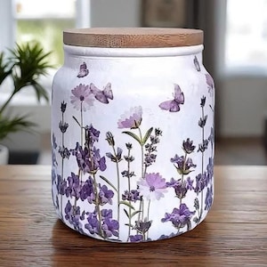 May include: White ceramic jar with a wooden lid, decorated with purple lavender flowers and butterflies. The jar is cylindrical and sits on a wooden surface. The design features a floral and butterfly theme.