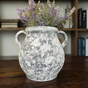 May include: A decorative, two-handled vase with a distressed, gray and white finish. The vase is filled with faux lavender stems, adding a touch of color. The vase is a decorative home accent.