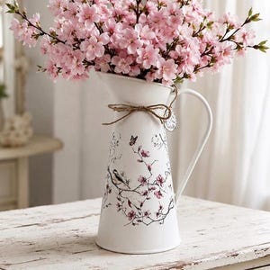May include: A white metal pitcher featuring a tapered body with a black and pink floral and bird illustration. A jute twine bow is tied around the neck, and the pitcher is filled with a large arrangement of light pink cherry blossom branches.