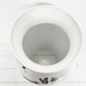 K&ouml;nnte beinhalten: Wei&szlig;e Keramikvase mit einem schwarz-wei&szlig;en Schmetterlings- und L&ouml;wenzahnmuster.