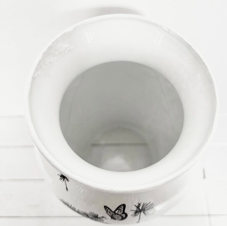 K&ouml;nnte beinhalten: Wei&szlig;e Keramikvase mit einem schwarz-wei&szlig;en Schmetterlings- und L&ouml;wenzahnmuster.