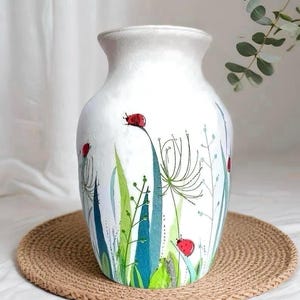 May include: White ceramic vase with a wide base and a slightly flared top. The vase is decorated with a hand-painted design of green and blue foliage, with red ladybugs scattered throughout. The vase sits on a round, woven, tan mat.