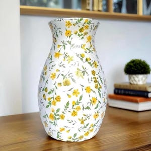 May include: White ceramic vase with a floral pattern of yellow flowers and green leaves. The vase has a wide base and a slightly flared top. The vase is a decorative item for the home.