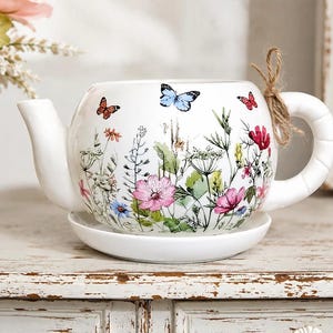 May include: A white ceramic teapot-shaped planter with a matching saucer, featuring a colorful botanical print of wildflowers, green foliage, and small butterflies. A piece of brown twine is tied around the handle of the white ceramic vessel.