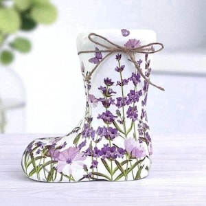 May include: A white ceramic boot-shaped vase decorated with purple lavender flowers and green leaves. The vase has a twine bow at the top. The design includes butterflies and a floral pattern, suitable for home decor.