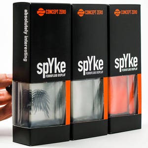 Könnte beinhalten: Drei schwarze Boxen mit orangenen Akzenten, jede mit einem durchsichtigen Behälter gefüllt mit einer schwarzen Flüssigkeit. Die Boxen sind mit "spyke FERROFLUID DISPLAY" und "CONCEPT ZERO" beschriftet. Der Text "absolutely interesting" ist auf der linken Box aufgedruckt.