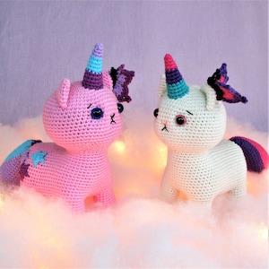 Op de afbeelding: Twee handgemaakte gehaakte eenhoornkattenknuffels. Eén is roze met een blauwe ster en de andere is wit. Beide hebben regenbooghoorns, paarse vlinderaccenten en staan tegen een zachte, wolkige achtergrond.