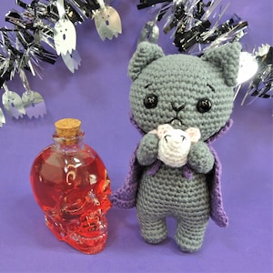 Puede incluir: Un gato de crochet gris vestido de vampiro sosteniendo un ratón de crochet blanco. El gato lleva una capa morada y tiene ojos negros. El gato está de pie sobre una superficie morada junto a una botella de vidrio transparente con forma de cráneo llena de líquido rojo.