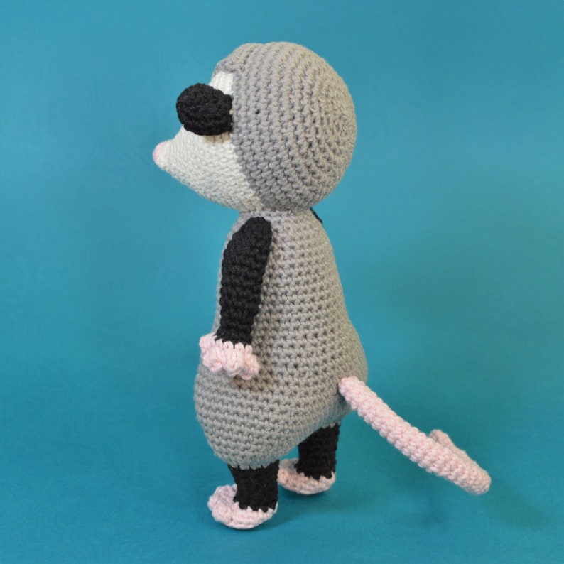 Pu&ograve; includere: Un opossum di uncinetto grigio con accenti neri e rosa. L'opossum &egrave; in piedi su uno sfondo blu.