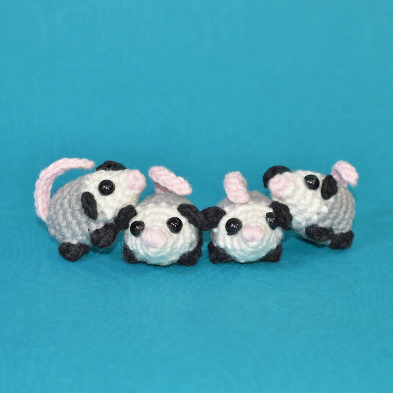 Pu&ograve; includere: Tre peluche di planeur dello zucchero all'uncinetto in grigio, bianco e rosa con occhi neri. I peluche sono disposti in fila su uno sfondo blu.